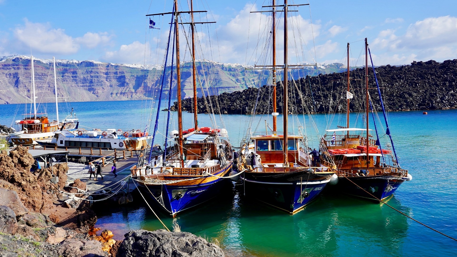 drewniane statki z masztami przycumowane do skalistego brzegu na tle klifów | kiedy najlepiej jechać do Grecji