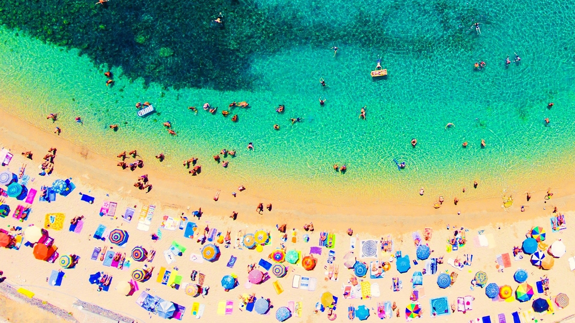 piaszczysta plaża z lotu ptaka z kolorowymi parasolami oblana turkusową wodą, w której pływają ludzie | kiedy najlepiej jechać do Grecji